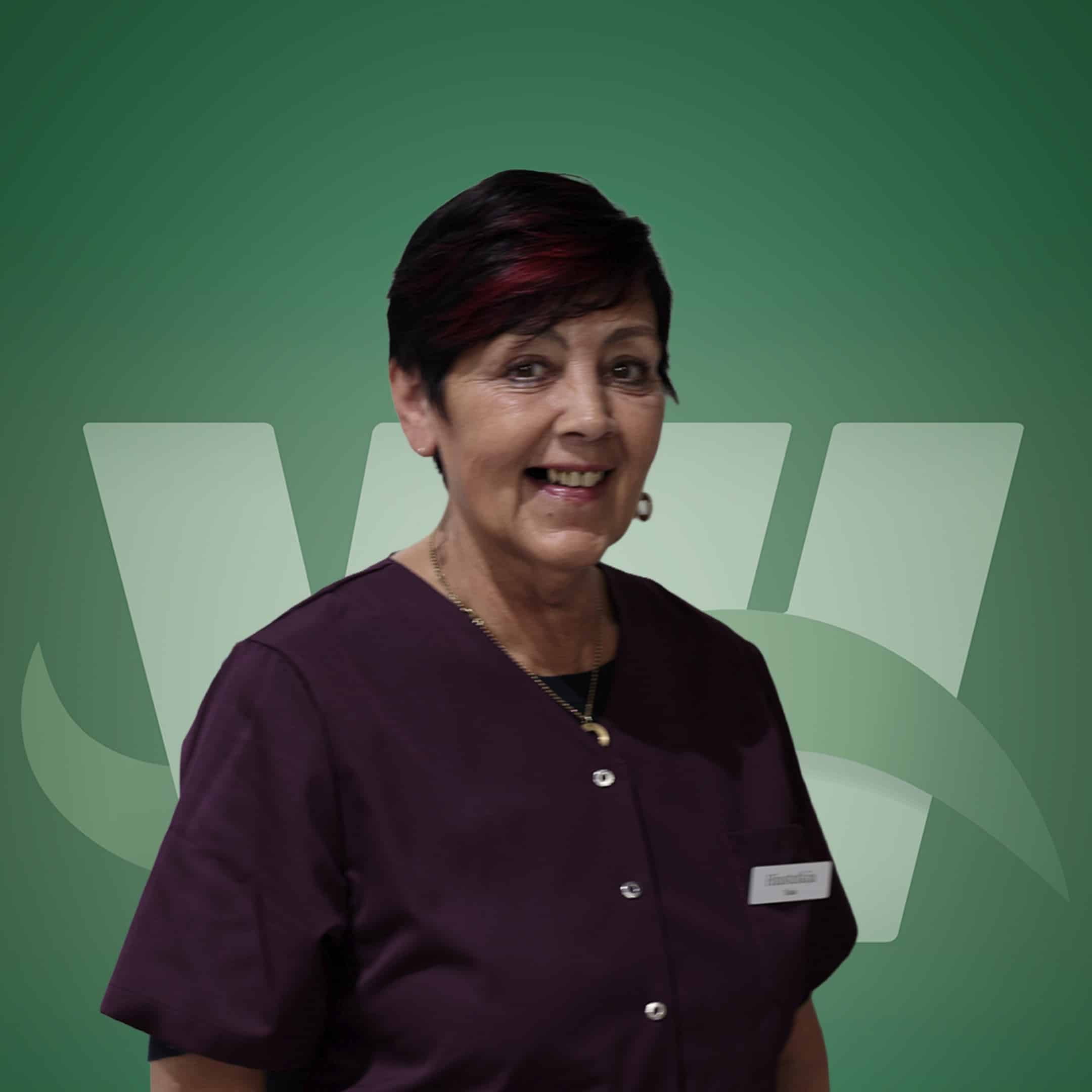 Smiling woman in a dark purple uniform with a name badge, standing against a green decorative background.